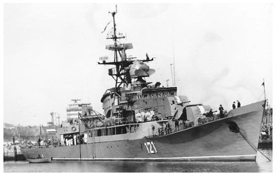 Das Küstenschutzschiff mit der Bordnummer 121 liegt im Stützpunkt an der Pier, aufgenommen schräg von vorn backbord mit Gittermast, Schornstein und 100-mm-Türmen